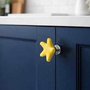 May include: A yellow starfish-shaped cabinet knob with a textured surface, mounted on a navy blue cabinet door. The knob has a chrome base. The background includes a white countertop and a white tile backsplash.