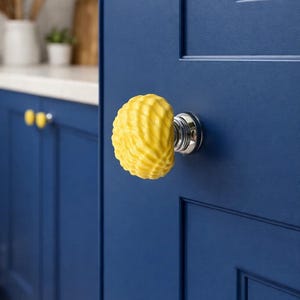 Może przedstawiać: Zbliżenie granatowych drzwi szafki z jasnoż&oacute;łtą, teksturowaną gałką w kształcie muszli. Gałka jest zamontowana na srebrnej podstawie. W tle widoczne są inne ż&oacute;łte gałki.