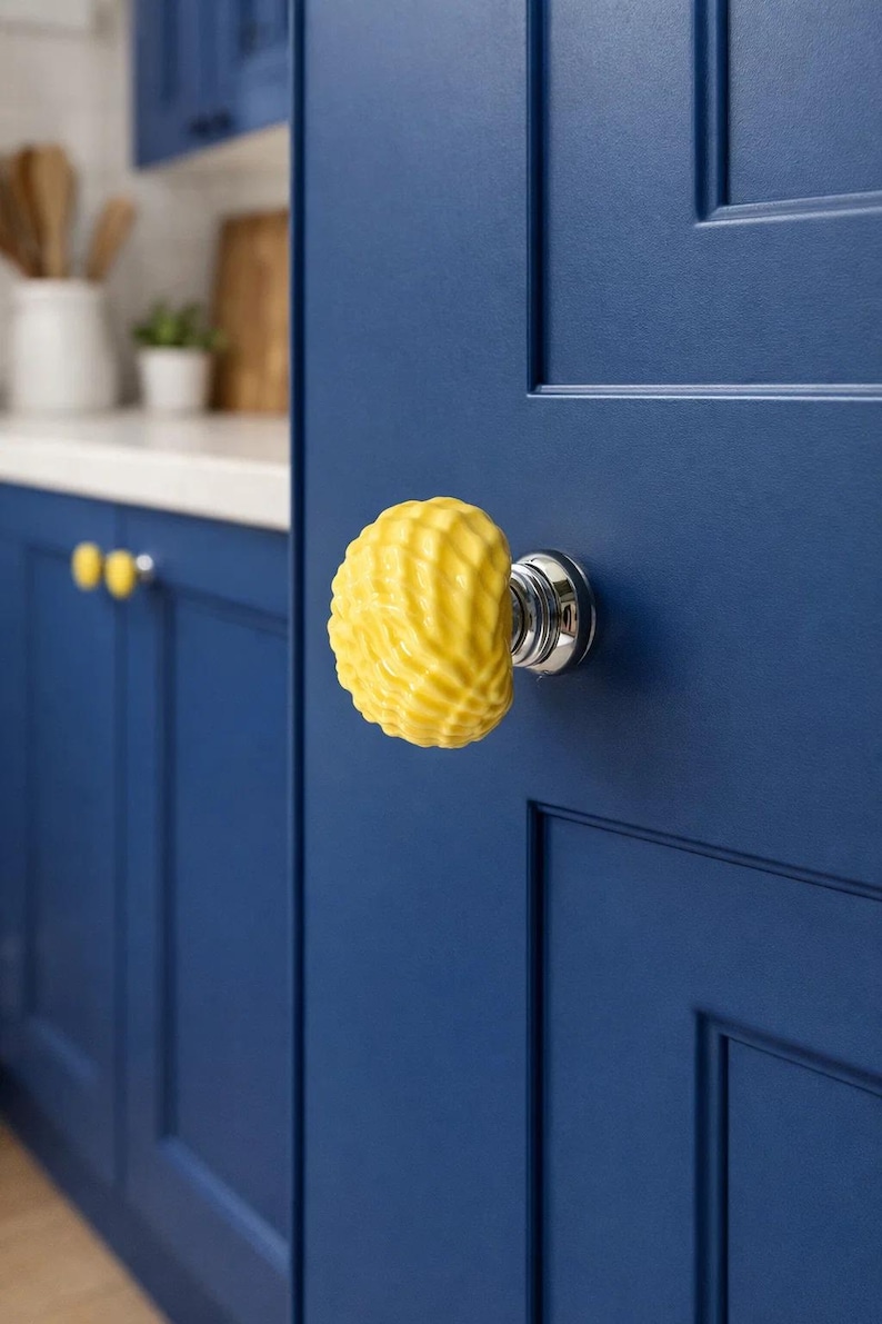 Może przedstawiać: Zbliżenie granatowych drzwi szafki z jasnoż&oacute;łtą, teksturowaną gałką w kształcie muszli. Gałka jest zamontowana na srebrnej podstawie. W tle widoczne są inne ż&oacute;łte gałki.