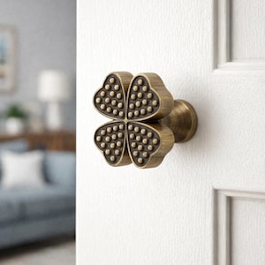 Op de afbeelding: Een bronzen deurknop in de vorm van een klavertjevier. Elk blad is getextureerd met kleine, gelijkmatig verdeelde stippen. De knop is gemonteerd op een witte deur, met een glimp van een kamer op de achtergrond.