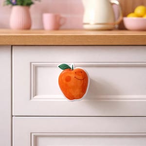 Könnte beinhalten: Ein orangefarbener, apfelförmiger Schrankknauf mit einem grünen Blatt und einem lächelnden Gesicht. Er ist an einer weißen Schranktür befestigt. Die Küche hat eine rosa geflieste Wand und eine Holz-Arbeitsplatte. Ein dekoratives Element.