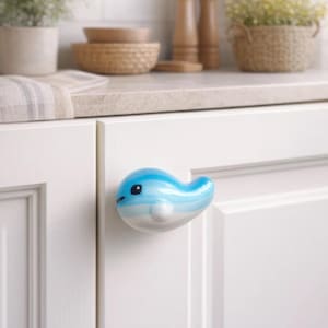 Puede incluir: Pomo de armario decorativo con forma de ballena azul y blanca, fijado a una puerta de armario blanca. La ballena tiene una cara sonriente y es un acento encantador para muebles de cocina o baño. El fondo incluye una encimera con objetos decorativos.