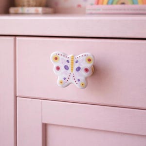 Könnte beinhalten: Rosa Schublade mit einem weißen, schmetterlingsförmigen Knauf. Der Knauf hat gelbe, rote und lila Akzente. Im Hintergrund ein Regal mit einem Regenbogen und einem Blumenkorb.