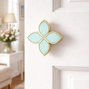 Könnte beinhalten: Dekorativer Türknauf in Form einer vierblättrigen Blume. Die Blütenblätter sind in einem sanften Aqua-Farbton gehalten und von goldfarbenem Metall umrandet. Der Knauf ist an einer weißen Tür befestigt, was auf einen Einrichtungsgegenstand hindeutet.