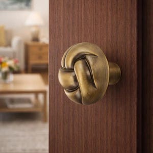 May include: A bronze-colored door knob in a knot design is mounted on a dark brown wooden door. The knob has a rounded shape with intricate detailing, suggesting a nautical or rustic theme. The background is blurred, showing a glimpse of a living room.