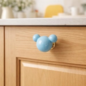 Könnte beinhalten: Hellblauer Keramik-Schrankknauf in Form eines Mäusekopfes, montiert an einer Holzschranktür. Der Knauf hat drei runde Kugeln und einen goldfarbenen Sockel. Die Schranktür ist aus hellem Holz gefertigt.