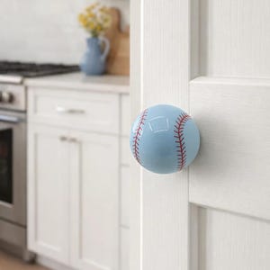 Könnte beinhalten: Hellblauer Türknauf im Baseball-Design mit roten Nähten, an einer weißen Tür befestigt. Der Knauf ist rund und scheint aus Keramik oder Glas zu sein. Im Hintergrund eine Küche mit weißen Schränken und einem blauen Krug.
