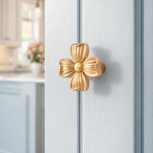 May include: A gold-colored, flower-shaped cabinet knob. The knob is mounted on a light blue door. The flower design has four petals with a central, rounded detail. The background shows a glimpse of a kitchen.