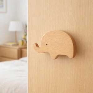 Könnte beinhalten: Ein hölzerner Türgriff in Elefantenform, montiert an einer hellfarbigen Holztür. Der Griff ist hellbraun mit einem kleinen, runden Augendetail. Der Rüssel des Elefanten ist nach unten gebogen. Der Hintergrund zeigt ein Schlafzimmer mit Bett und Nachttisch.
