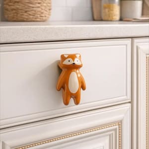 May include: A ceramic fox-shaped cabinet knob in orange and white. The knob is fixed to a white cabinet door. The fox has a white belly and face, with black eyes and a small black nose. The cabinet door has a gold trim.