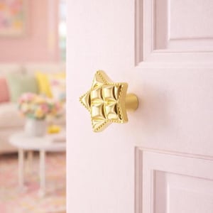 Pode incluir: Um puxador de porta dourado em forma de estrela com um design acolchoado está montado numa porta rosa claro. O puxador tem uma base arredondada e uma superfície texturizada. O fundo mostra um vislumbre de uma sala com um sofá branco e uma mesa redonda.