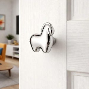 May include: A polished chrome door knob in the shape of a horse. The horse-shaped knob is mounted on a white door. The knob has a smooth, reflective surface and is designed with rounded features.