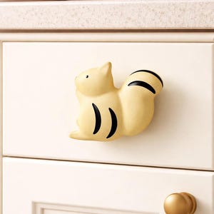 May include: A light yellow cat-shaped cabinet knob with black stripes on the tail and body. The knob is mounted on a white cabinet door. A gold-colored knob is visible below the cat knob.