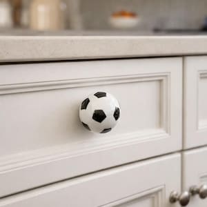 May include: White cabinet door with a soccer ball-shaped knob. The knob is white with black pentagon shapes. The cabinet is part of a white kitchen set. The background is blurred, showing a countertop and a vase of flowers.