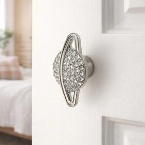 May include: A silver door knob with a decorative design. The knob has a central oval shape covered in small, sparkling crystals, framed by a curved, open structure. It is mounted on a white door.