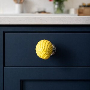 Może przedstawiać: Jasnoż&oacute;łta gałka do szafki w kształcie muszli z srebrną podstawą jest zamontowana na ciemnoniebieskich drzwiach szafki. Gałka dodaje dekoracyjnego akcentu do szafek kuchennych. W tle widać blat i wazon z kwiatami.