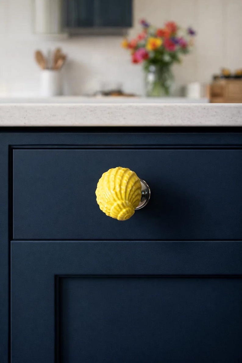 Może przedstawiać: Jasnoż&oacute;łta gałka do szafki w kształcie muszli z srebrną podstawą jest zamontowana na ciemnoniebieskich drzwiach szafki. Gałka dodaje dekoracyjnego akcentu do szafek kuchennych. W tle widać blat i wazon z kwiatami.