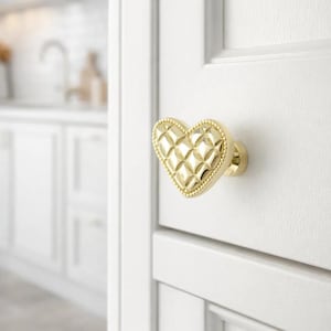 Puede incluir: Pomo de armario dorado en forma de corazón con diseño acolchado y borde de cuentas. El pomo está montado en una puerta de armario blanca. El fondo muestra un entorno de cocina borroso.