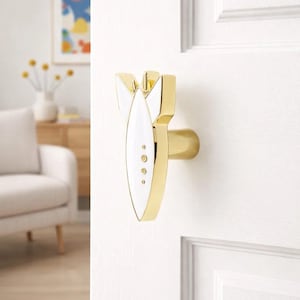 May include: A gold-toned door handle with a white and gold design. The handle has a unique shape, with a central white oval and three small circles. The handle is mounted on a white door.