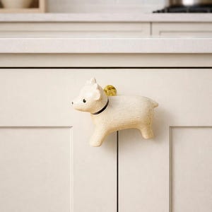 Puede incluir: Pomo de armario en forma de cerdo de color crema, con un collar negro y un acento dorado. El pomo decorativo está montado en una puerta de armario blanca. El entorno de la cocina incluye una encimera blanca y una olla de acero inoxidable.