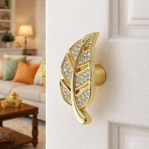 Peut inclure: Un bouton de porte doré en forme de feuille orné de cristaux transparents incrustés. Le bouton est monté sur une porte blanche. Le design de la feuille présente une veine centrale et des bords détaillés.