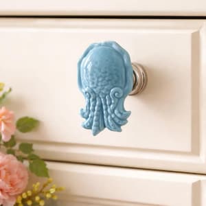 Könnte beinhalten: Hellblauer Keramik-Schrankknauf in Oktopusform mit silberfarbenem Metallsockel. Der Knauf ist an einer weißen Schranktür befestigt. Das Oktopus-Design weist strukturierte Details und eine glänzende Oberfläche auf.