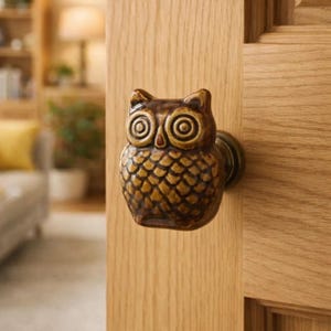 Może przedstawiać: Ozdobna gałka do drzwi w kształcie sowy, wykonana z ceramiki. Gałka jest brązowo-beżowa z szczegółowym wzorem, w tym teksturowanymi piórami i dużymi, okrągłymi oczami. Zamontowana na jasnych drewnianych drzwiach.
