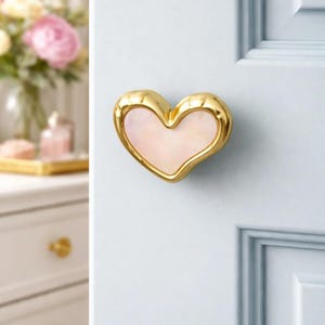 May include: A heart-shaped door knob with a gold finish and a pearlescent pink center. The knob is mounted on a light blue door. The design is elegant and decorative, suitable for home decor.