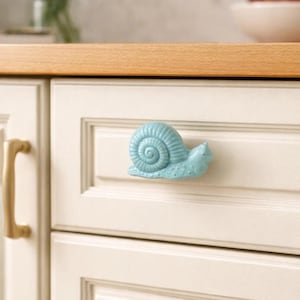 May include: A light blue ceramic snail-shaped cabinet knob. The knob is mounted on a white cabinet door with a gold-colored handle. The snail has a detailed shell and a textured body. The kitchen cabinet has a wooden countertop.