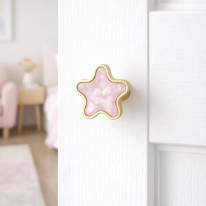 Può includere: Pomello per armadio a forma di stella con un centro rosa marmorizzato e un bordo dorato. Il pomello è montato su una porta bianca. Il pomello è largo circa 7.6 cm.