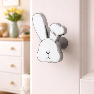 May include: A silver and white bunny-shaped door knob. The knob is mounted on a white door. The bunny has a white face and ears with a silver outline. The door knob is a decorative element for a child's room.