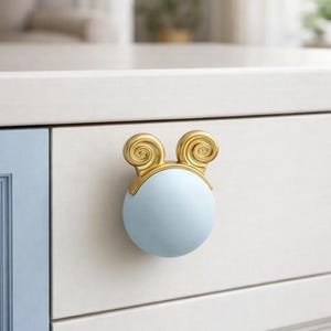 Puede incluir: Un pomo de armario azul claro con una parte superior decorativa dorada. El pomo es redondo y tiene un diseño de remolino en la parte superior. El pomo está montado en una puerta de armario blanca.