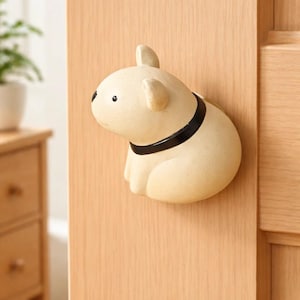 Puede incluir: Un tope de puerta beige con forma de perro y collar negro, montado en una puerta de madera clara. El tope tiene orejas pequeñas y redondeadas y un acabado liso. El perro está sentado.