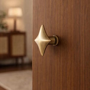 Może przedstawiać: Mosiężna klamka do drzwi w kształcie gwiazdy jest zamontowana na drewnianych drzwiach. Klamka ma geometryczny kształt z czterema punktami. Drzwi są w ciepłym brązowym kolorze, a tło jest lekko rozmyte.