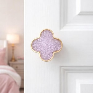 May include: A four-leaf clover-shaped door knob with a gold-colored frame and a light purple, crystal-like center. The knob is mounted on a white door. The background shows a bedroom with a lamp and bed.