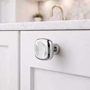 Puede incluir: Un pomo de armario cromado pulido con una cara cuadrada y redondeada. El pomo está montado en una puerta de armario blanca, que refleja la cocina circundante. El herraje añade un toque moderno a la carpintería.