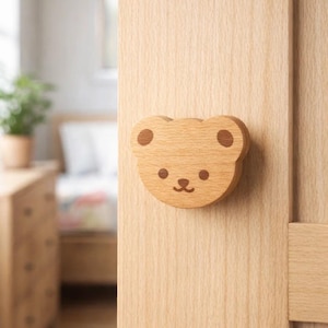 May include: A wooden cabinet knob in the shape of a bear's face. The knob is made of light-colored wood with a smooth finish. The bear's face has two dark brown circles for eyes, a small nose, and a smiling mouth. The knob is attached to a light-colored wooden cabinet door.