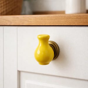 May include: A bright yellow ceramic cabinet knob with a vase-like shape. The knob is mounted on a brass-colored base and attached to a white cabinet door. The knob adds a pop of color to the neutral-toned kitchen.