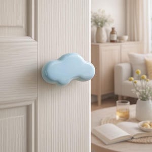 May include: A pale blue cloud-shaped door handle. It's fixed to a white wooden door. The background reveals a living room with a round wooden table, a sofa, and decorative items.