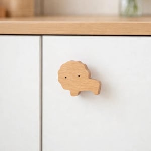 May include: A light-colored wooden cabinet knob in the shape of a lion's head. The knob is attached to a white cabinet door. The cabinet is part of a light-colored wooden cabinet. The knob has two small, dark dots for eyes.