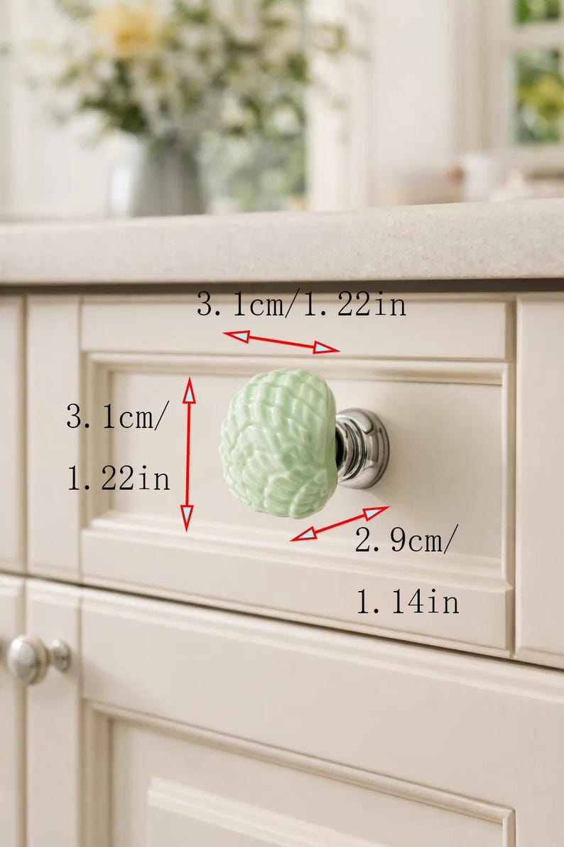 Może przedstawiać: Jasnozielony ceramiczny uchwyt do szafki z motywem muszli, zamontowany na białych drzwiach szafki. Uchwyt ma średnicę 3,1 cm i długość 2,9 cm. Drzwi są częścią kuchni lub łazienki.