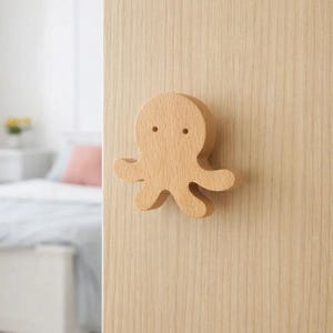 Puede incluir: Un tirador o pomo de puerta de madera con forma de pulpo, montado en una puerta de madera de color claro. El pulpo tiene un diseño sencillo y de dibujos animados con dos pequeños agujeros para los ojos y cuatro brazos. La puerta y el pomo están en un dormitorio.