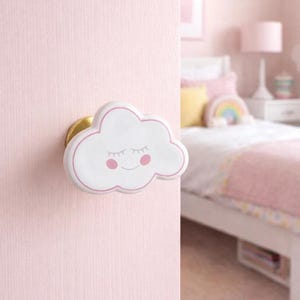 Puede incluir: Pomo de puerta blanco en forma de nube con detalles rosas y base dorada. El pomo, con ojos cerrados y mejillas rosadas, está montado en una puerta rosa, ideal para un dormitorio infantil.