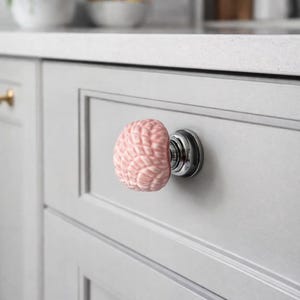 Może przedstawiać: R&oacute;żowy ceramiczny uchwyt do szafki w kształcie m&oacute;zgu, zamontowany na srebrnej podstawie. Uchwyt jest przymocowany do jasnoszarych drzwi szafki. Tło przedstawia biały blat i fragment kuchni.