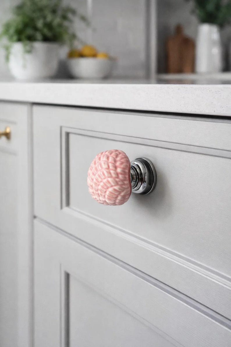 Może przedstawiać: R&oacute;żowy ceramiczny uchwyt do szafki w kształcie m&oacute;zgu, zamontowany na srebrnej podstawie. Uchwyt jest przymocowany do jasnoszarych drzwi szafki. Tło przedstawia biały blat i fragment kuchni.