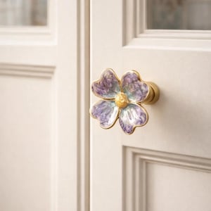 May include: A decorative door knob in the shape of a four-petal flower. The flower has a gold-colored rim and a center with a yellow hue. The petals are a mix of purple and white. The knob is mounted on a white door.