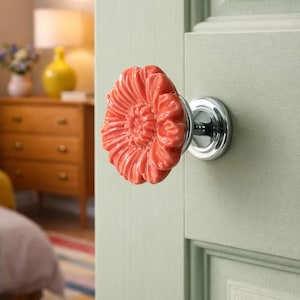 May include: A coral-colored, flower-shaped ceramic door knob with a chrome base is attached to a light green door. The knob has detailed petal-like ridges. The background shows a glimpse of a bedroom with a dresser and a lamp.
