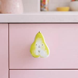 Könnte beinhalten: Ein skurriler, birnenförmiger Schrankknauf, gelb und weiß bemalt mit einem lächelnden Gesicht. Der Knauf ist an einer rosa Schranktür befestigt und verleiht der Küchendekoration eine verspielte Note.