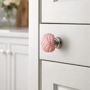 Może przedstawiać: R&oacute;żowy ceramiczny uchwyt do szafki o fakturze przypominającej m&oacute;zg, zamontowany na białych drzwiach szafki z srebrną podstawą. Uchwyt dodaje unikalnego charakteru szafkom kuchennym lub łazienkowym. Tło pokazuje fragment kuchni.