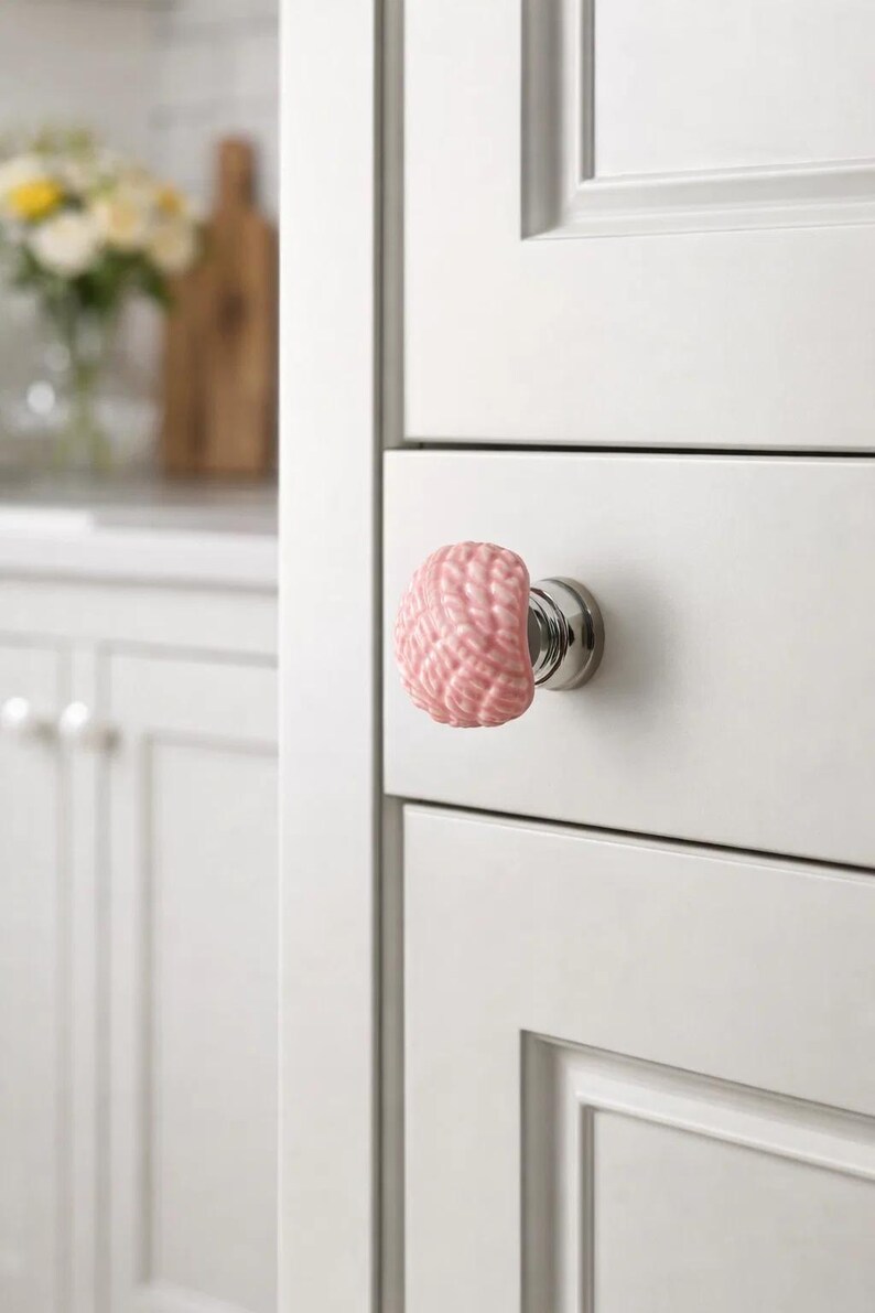 Może przedstawiać: R&oacute;żowy ceramiczny uchwyt do szafki o fakturze przypominającej m&oacute;zg, zamontowany na białych drzwiach szafki z srebrną podstawą. Uchwyt dodaje unikalnego charakteru szafkom kuchennym lub łazienkowym. Tło pokazuje fragment kuchni.